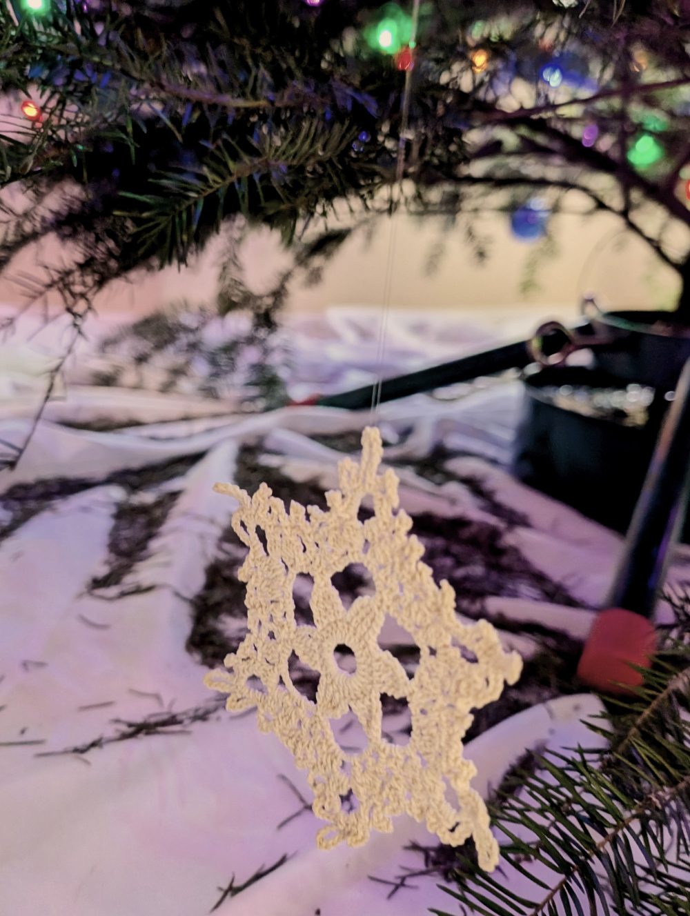 A handmade snow flake hangs from a Christmas tree, lights glowing in the background.